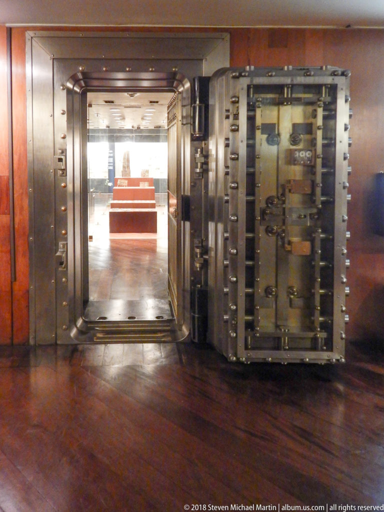 Open massive steel bank-vault door with rows of bolts leading into a museum gallery with display plinths.