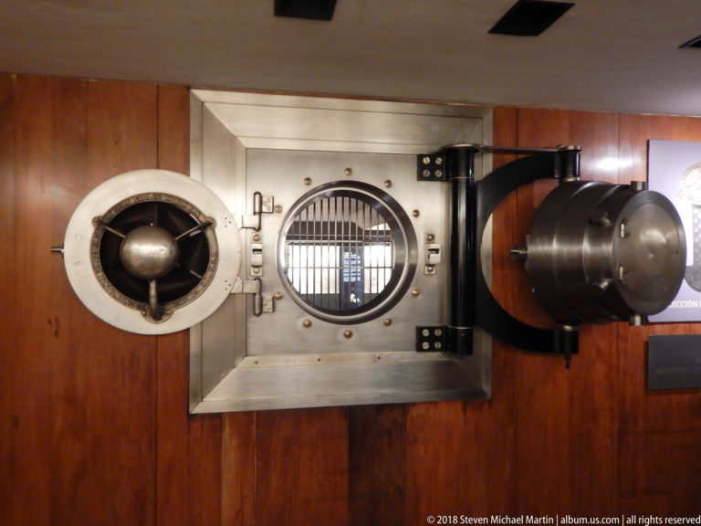 Heavy circular steel bank-vault mechanism beside a barred round opening set into a wood-paneled wall.