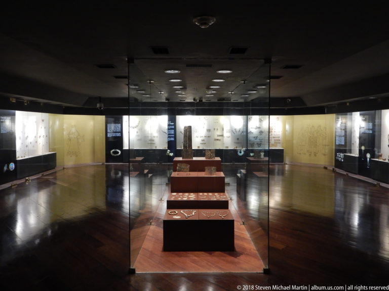 Dark museum gallery with illuminated wall cases, central display plinths, glass partitions, and polished wood floor.