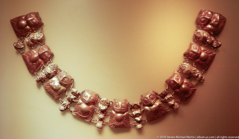 Curving necklace composed of repeated reddish metal human-figure pendants linked by smaller bright decorative elements.