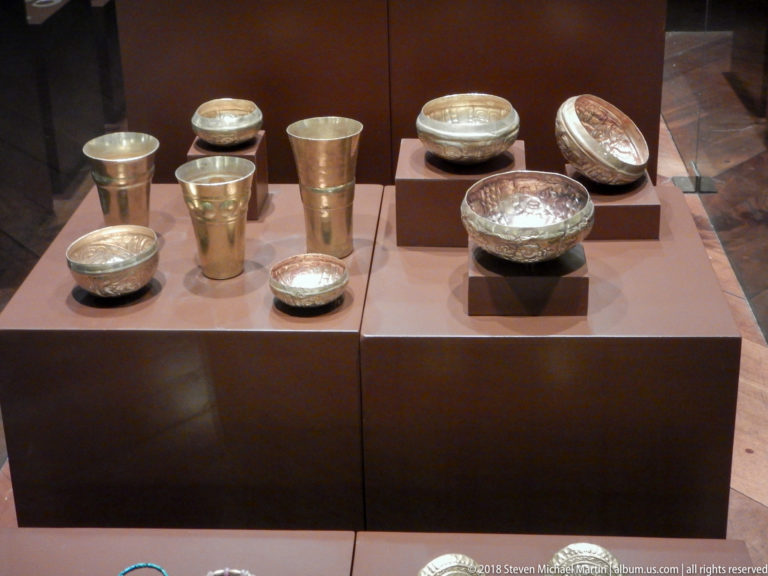 Silver-colored cups, beakers, bowls, and decorated vessels arranged on brown museum display plinths.