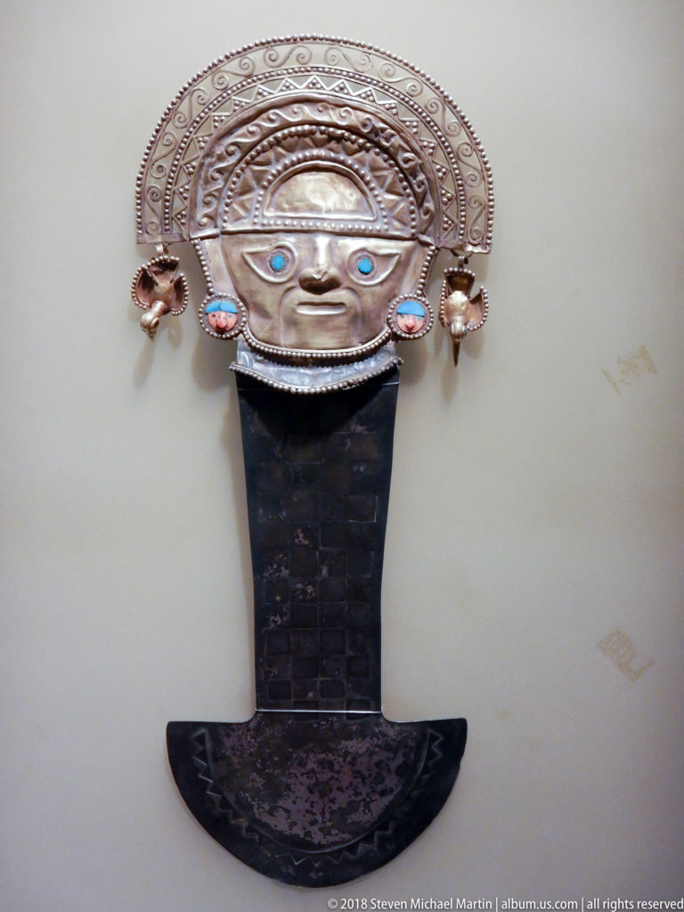 Anthropomorphic metal ornament with arched embossed headdress, blue-green inlaid eyes, side pendants, and broad dark lower element.