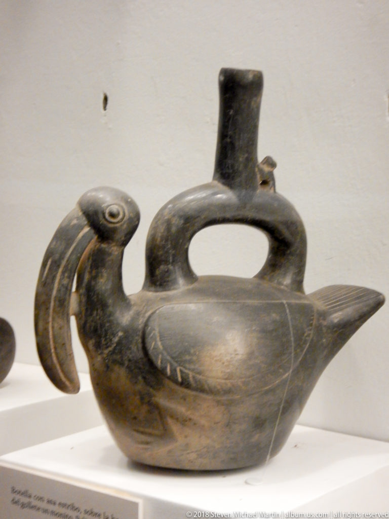 Dark ceramic bird-shaped vessel with curved neck, incised wing, handle-like opening, and upright spout.