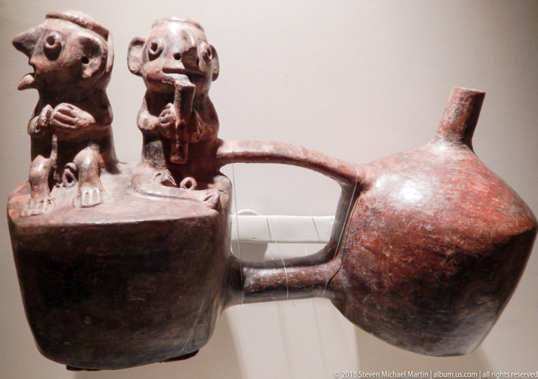 Reddish-brown pre-Hispanic ceramic vessel with two modeled human figures, rounded body, bridge tube, and upright spout.