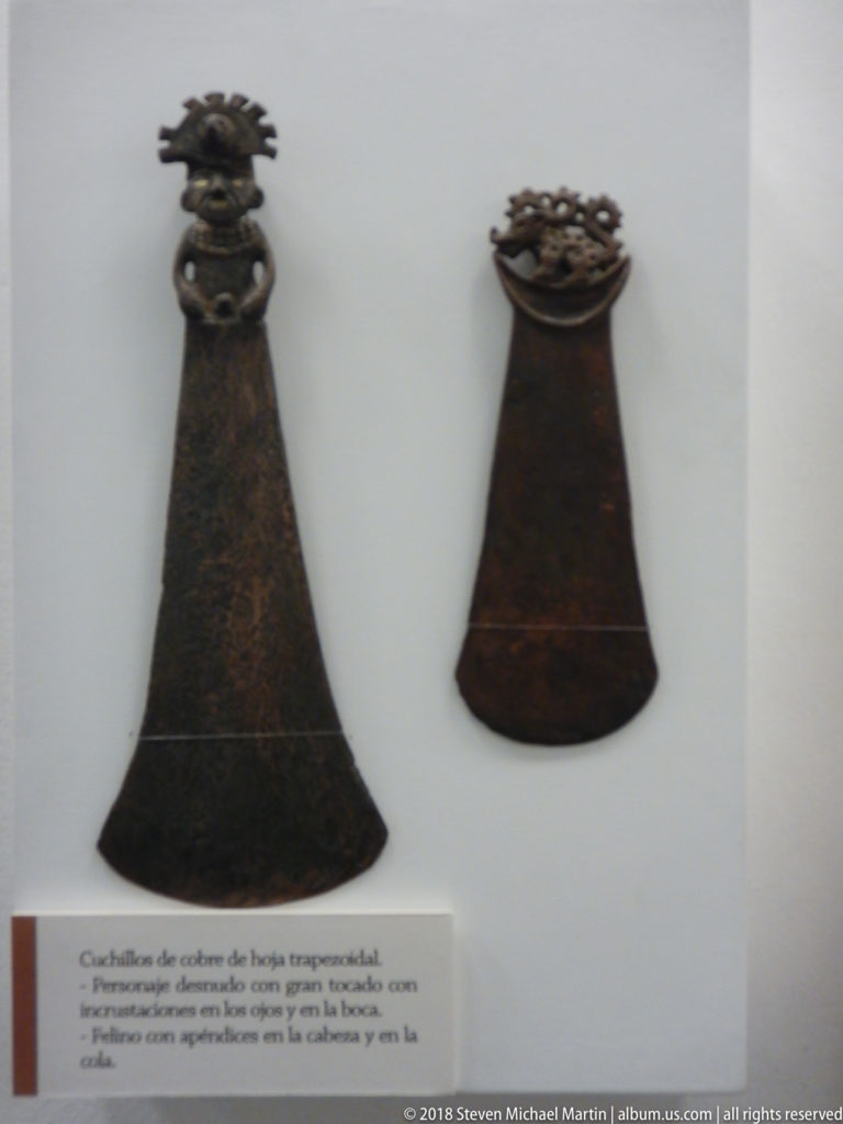 Two dark trapezoidal copper knives or blades displayed vertically above a Spanish museum label.