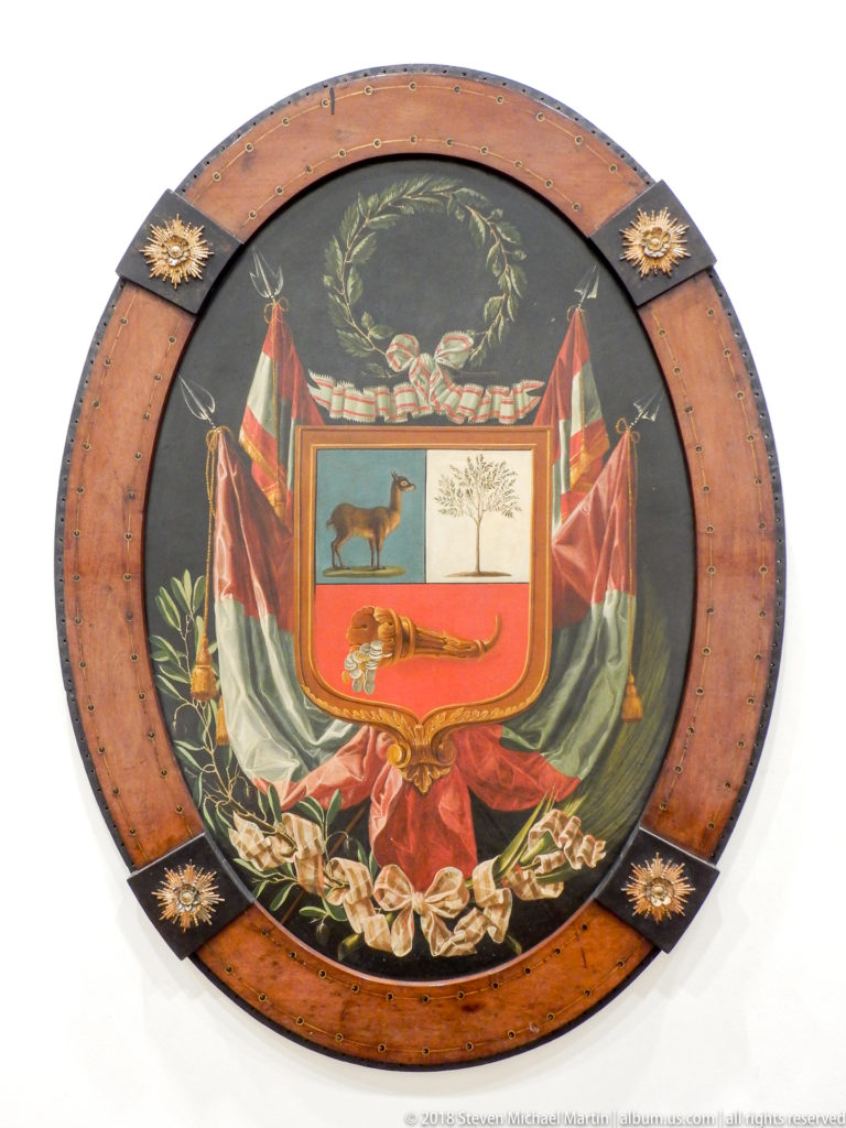 Decorative oval plaque with Peru’s coat of arms, national flags, wreath, vicuña, cinchona tree, and cornucopia.