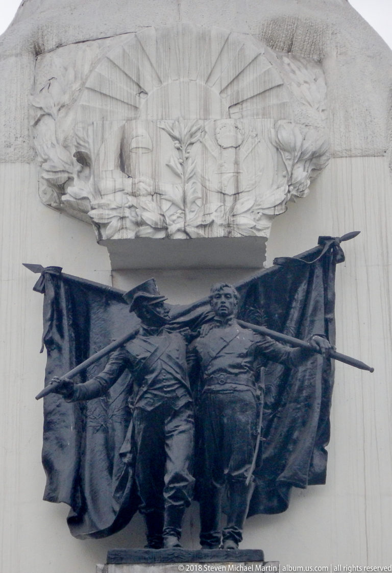 Dark bronze relief of advancing armed figures and flags mounted beneath a carved pale stone canopy.