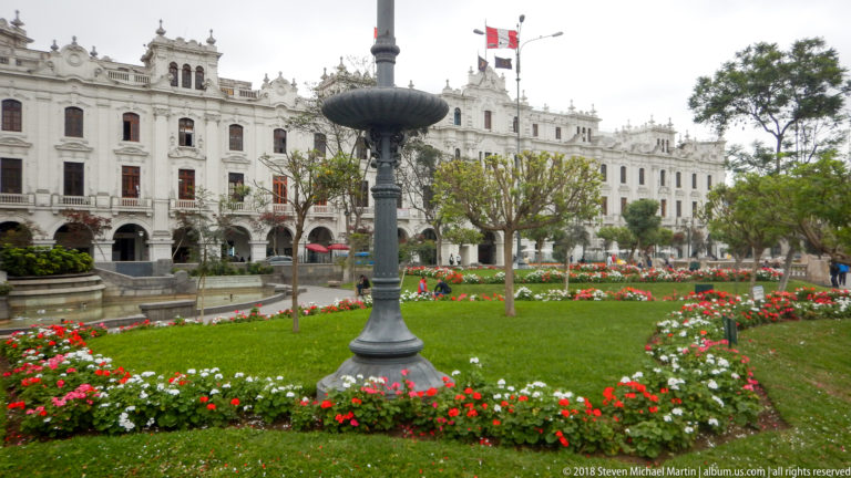 Flower beds and lawn in a historic Lima square, with an ornate iron fixture and white civic buildings beyond.