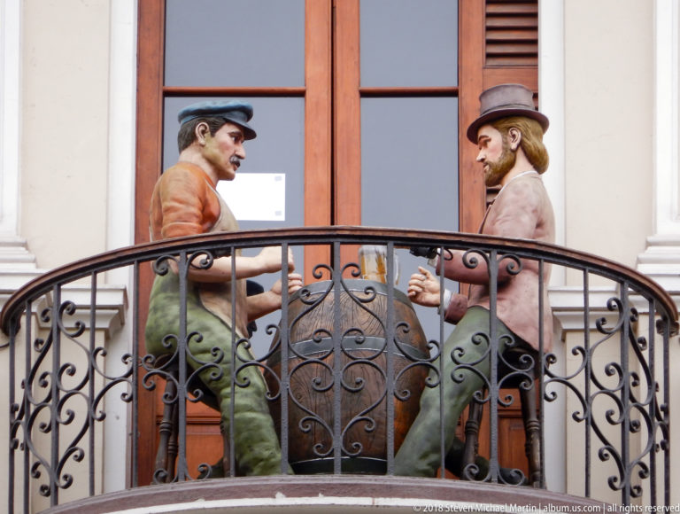 Two life-size decorative male figures beside a large vessel on a curved wrought-iron balcony.