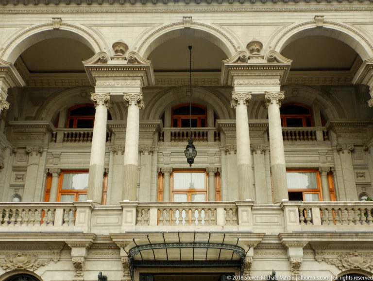 Upper façade with paired pale columns, rounded arches, balustrade, wooden windows, and a glass entrance canopy.