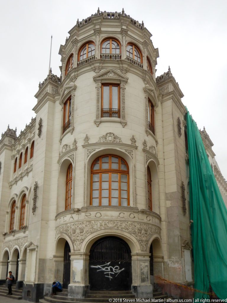 Curved cream-colored historic façade with arched wooden windows, carved ornament, and green construction sheeting at right.