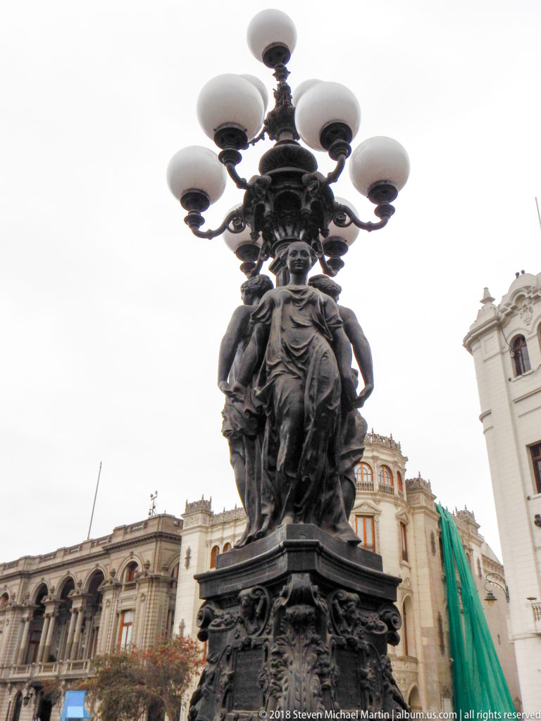 Low-angle view of an ornate dark metal street lamp with sculpted figures and multiple white globes in Lima.