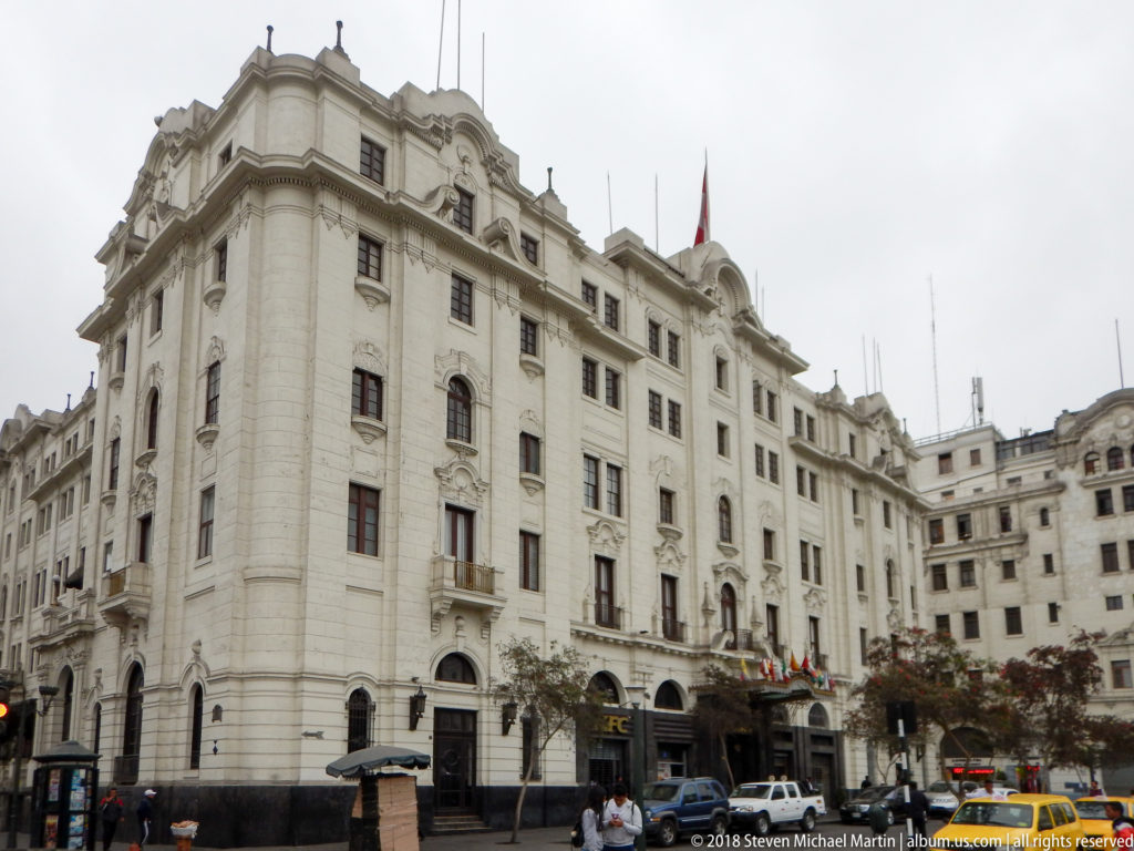Large white historic civic building viewed from a street corner with pedestrians, traffic, and a Peruvian flag.