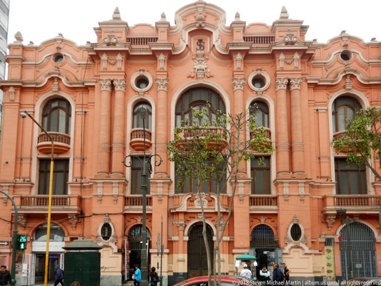Salmon-colored ornate civic façade with columns, arched windows, balconies, crest, street trees, and pedestrians.