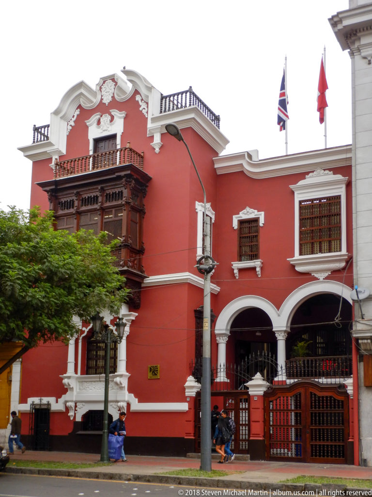 Red historic building with white trim, projecting carved wooden balcony, flags, and gated street entrance in Lima.