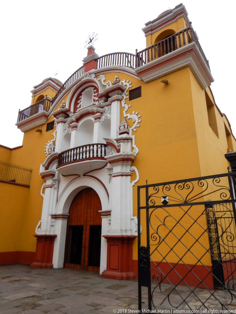 Oblique view of an ornate white portal on a yellow façade with a black wrought-iron gate in the foreground.