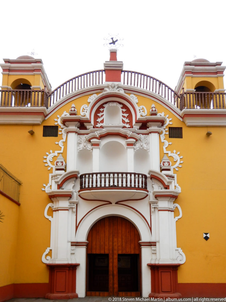 Frontal view of an ornate white and red portal and balcony on a mustard-yellow historic building in Lima.