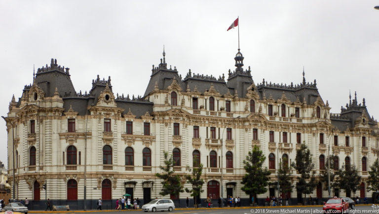 Large ornate government building with steep dark roofs, towers, red-framed windows, and a Peruvian flag in central Lima.