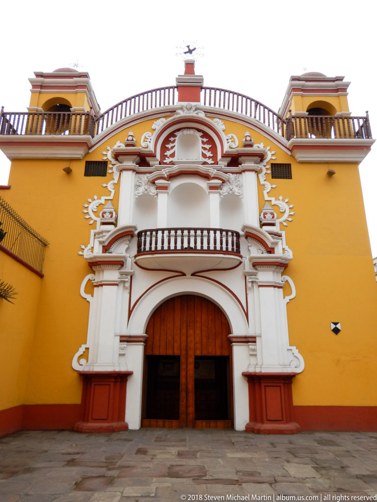 Ornate white and red portal with wooden doors and balcony set into a mustard-yellow historic façade in Lima.