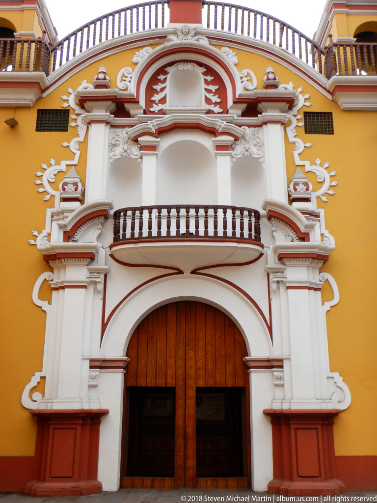Frontal yellow church façade with large wooden doors and an elaborate white-and-red portal rising to a curved balcony and upper ornament.