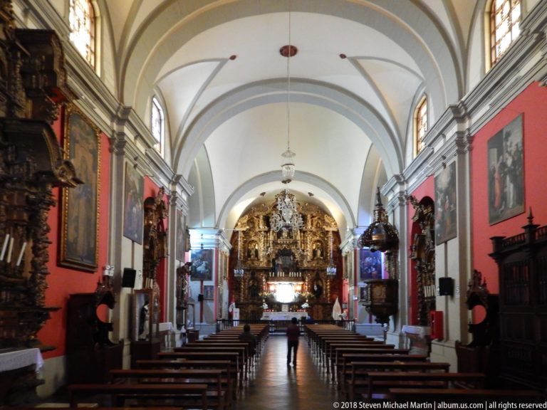 Oblique view down a church nave with dark pews, red walls, paintings, side altars, vaulted ceiling, and gilded sanctuary.