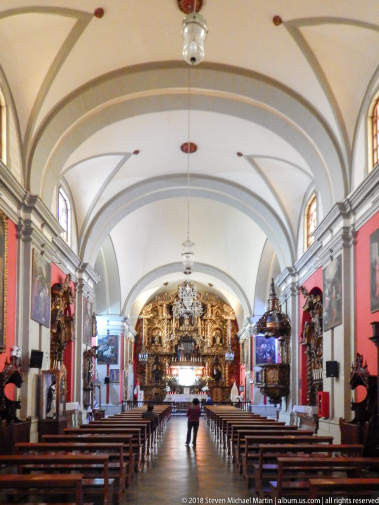 Long church nave with wooden pews, pale vaulted ceiling, red walls, side altars, and gilded sanctuary at the far end.