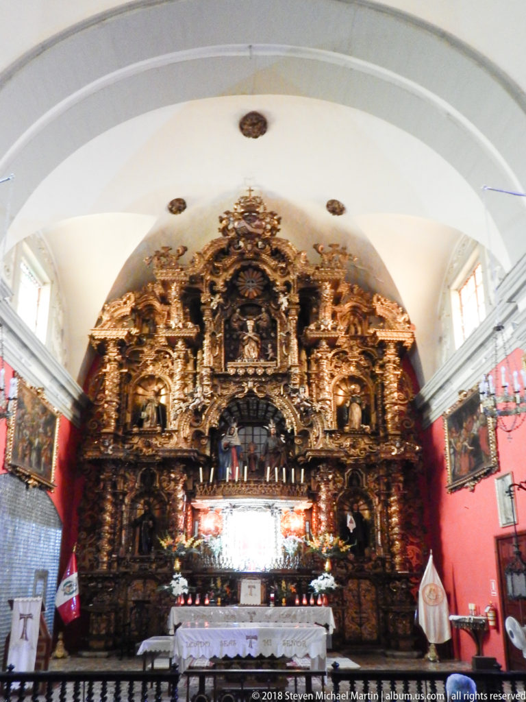 Monumental gilded retablo and altar beneath a white vaulted ceiling, with red side walls and religious paintings.