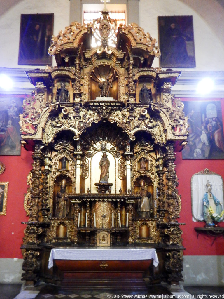 Gilded church retablo with twisted columns, central devotional figure, altar table, red walls, and framed paintings.
