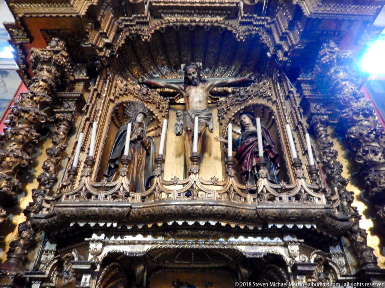 Crucified Christ with attendant figures, candles, and gilded carved ornament filling a richly decorated church retablo.