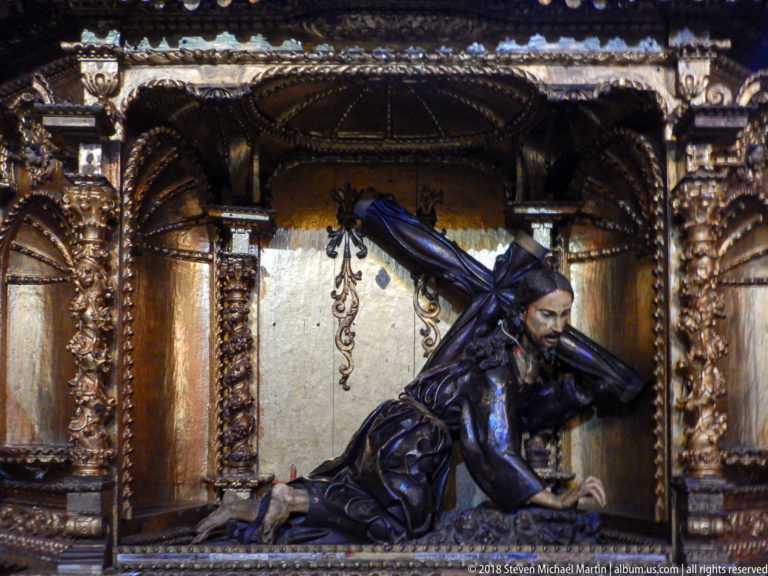 Polychrome reclining Christ figure beneath a dark cross inside a gilded niche framed by carved columns and dense ornament.