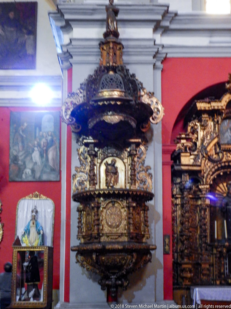 Gilded carved altarpieces line a red church wall with devotional paintings and images between the richly ornamented structures.