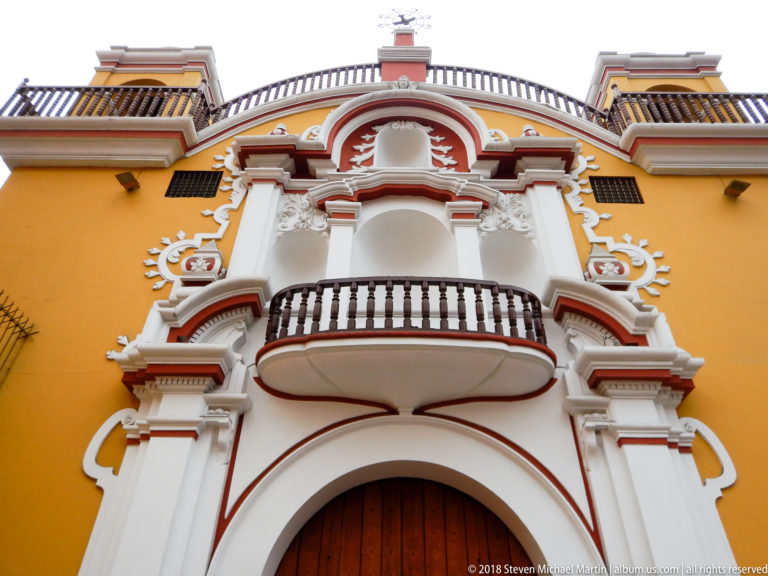 Yellow façade with elaborate white-and-red church portal, curved balcony, large wooden doorway, scrollwork, and crest-like upper ornament.