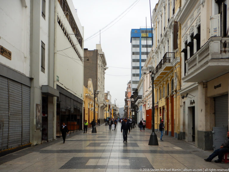 Pedestrians walk along a paved historic-center street lined with mixed commercial façades, balconies, and a modern tower in the distance.