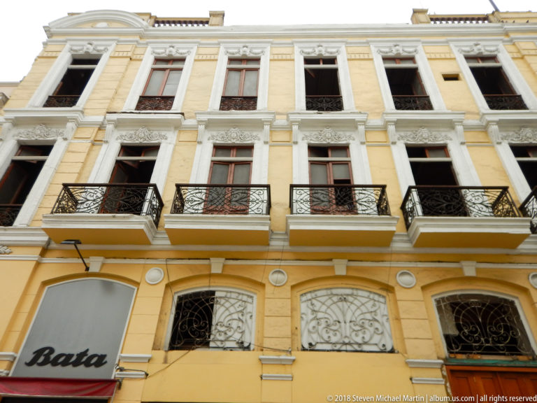 Yellow multi-story building with white trim, tall windows, black wrought-iron balconies, and a modern storefront at street level.