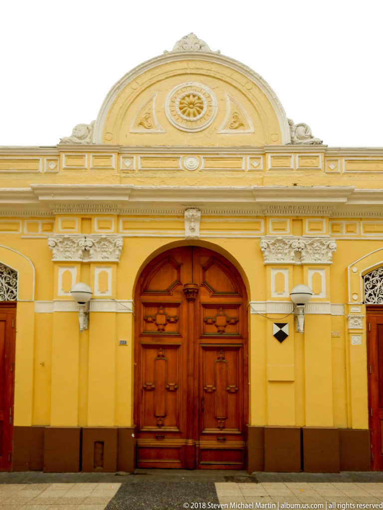 Large paneled wooden double door centered in a mustard-yellow façade with white classical moldings and a semicircular pediment.