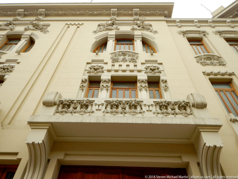 Upward view of a cream historic façade with carved balcony, floral reliefs, curved upper windows, and layered cornices.