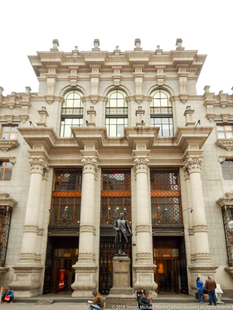 Monumental stone civic façade with tall columns, large gridded windows, central bronze statue, entrances, and pedestrians.