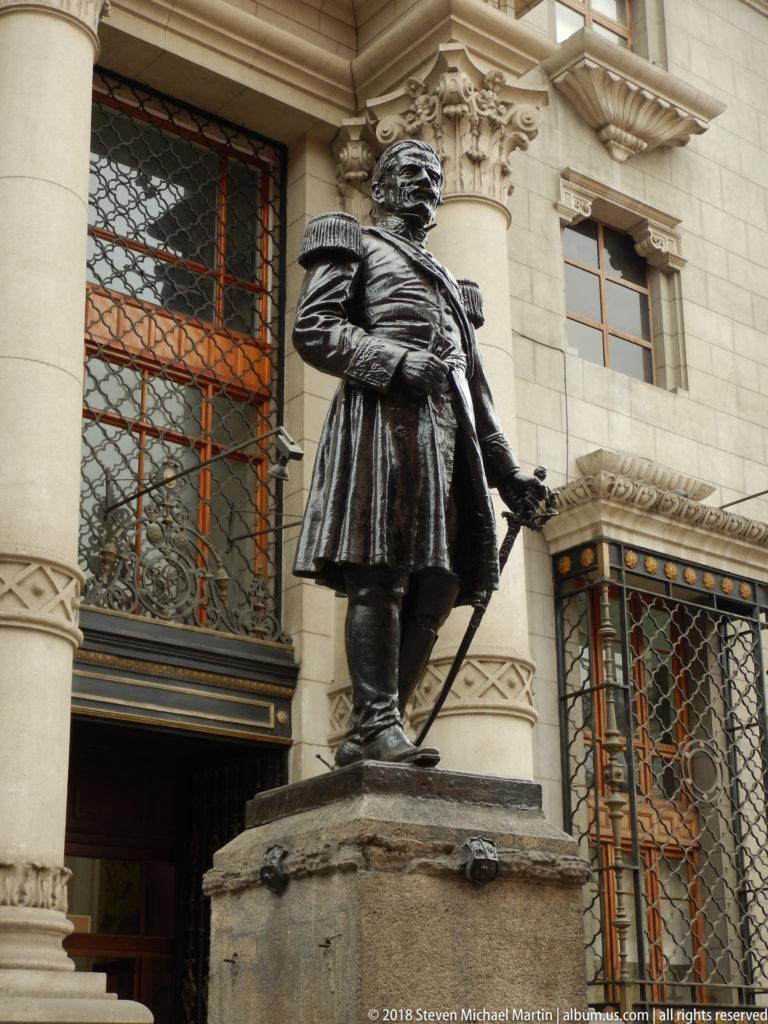 Bronze statue of a man in historical dress on a stone pedestal before a monumental building with gridded windows and carved columns.