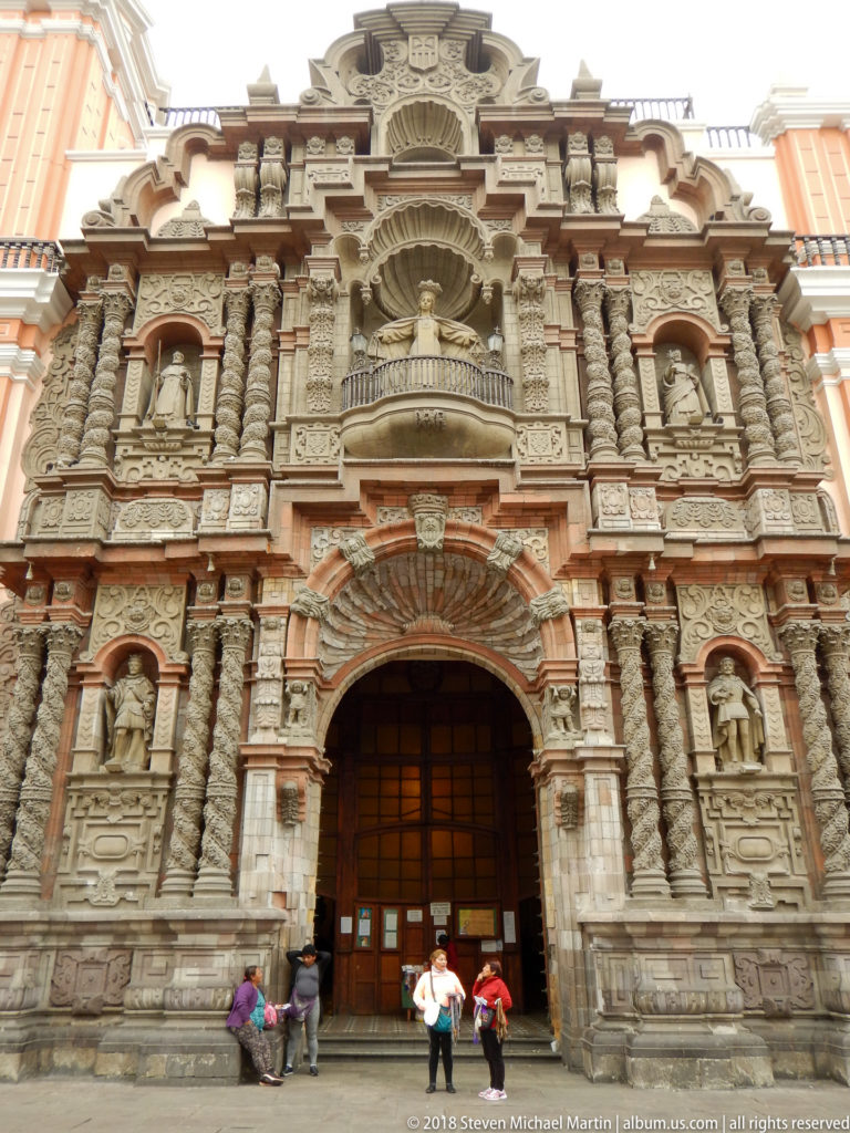 Frontal view of an ornate Baroque church portal with twisted columns, saint sculptures, shell niches, and visitors at the entrance.