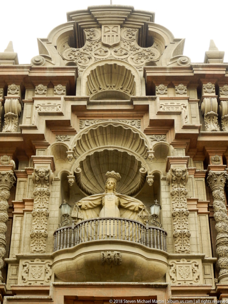 Upward view of an ornate stone church façade with carved columns, shell niches, reliefs, and a central balcony with sculpture.