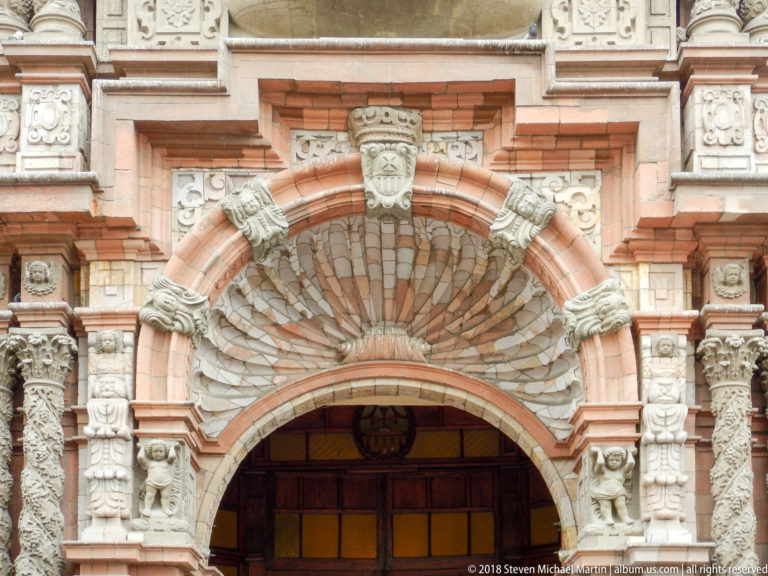 Close view of an ornate church doorway with reddish masonry, carved stone figures, columns, masks, and a fan-shaped arch.