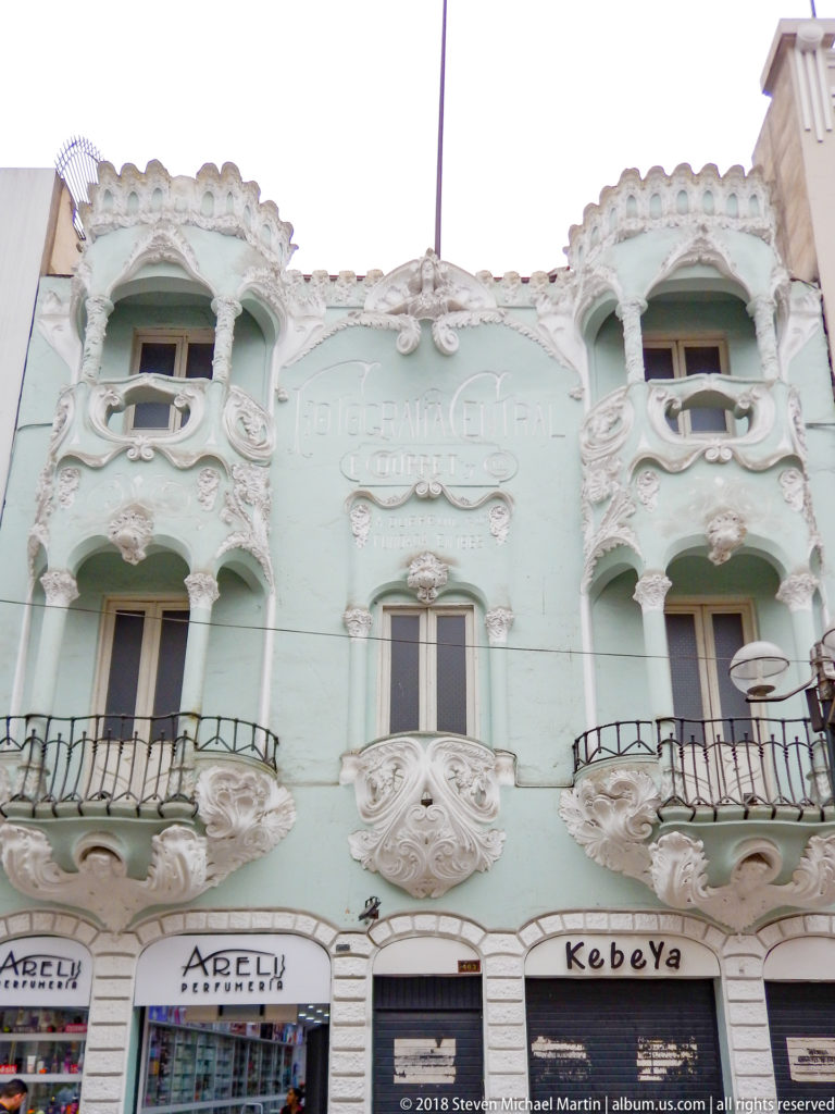 Pale green façade with elaborate white scrollwork, sculptural figures, curved balconies, and shopfronts at street level.