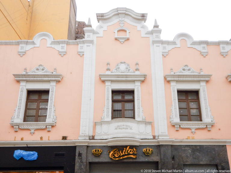 Pastel-pink upper façade with ornate white window surrounds, curving parapet, three wooden windows, and storefronts below.