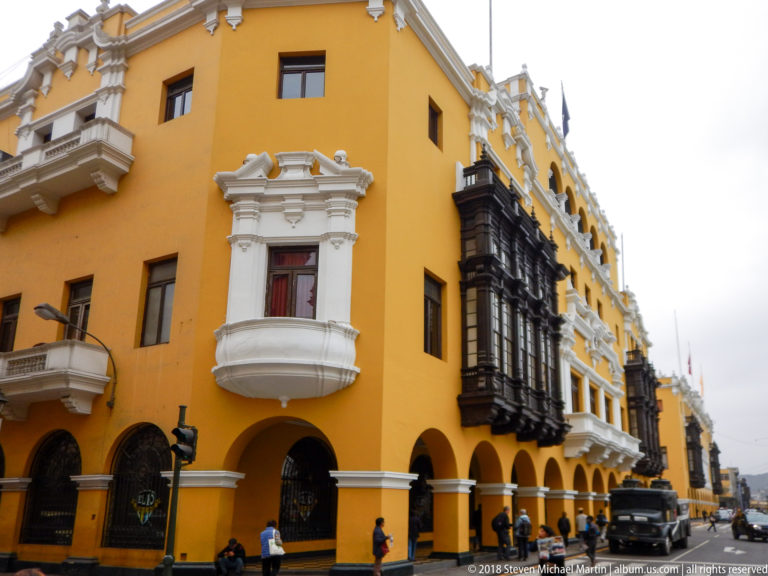 Yellow corner building with ground-floor arches, white ornamental trim, dark enclosed wooden balconies, pedestrians, and a receding street.