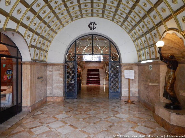 Arched entrance hall with patterned vaulted ceiling, stone walls, decorative iron gates, sculpture, and a staircase beyond.