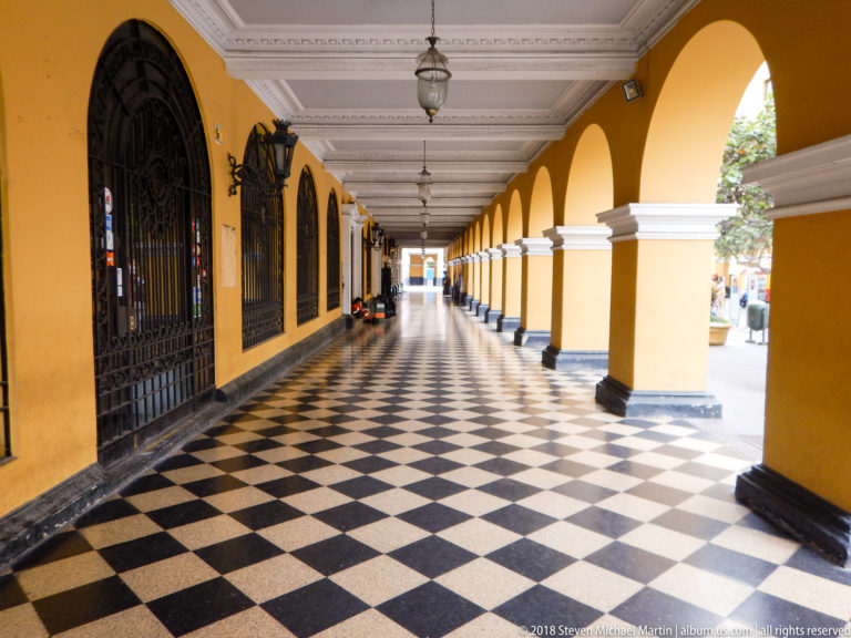 Long yellow arcade with checkerboard floor, white capitals, iron grilles, hanging lanterns, and bright courtyard openings to the right.