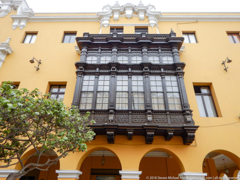 Large dark carved-wood enclosed balcony projecting from a yellow façade above arches and a leafy courtyard tree.