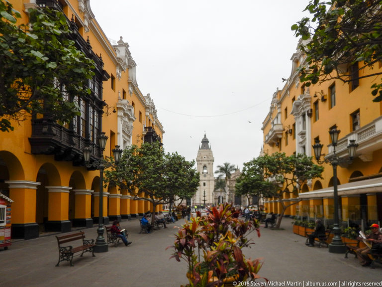 Yellow arcaded buildings and balconies frame a pedestrian courtyard with benches, plantings, and a pale tower in the distance.
