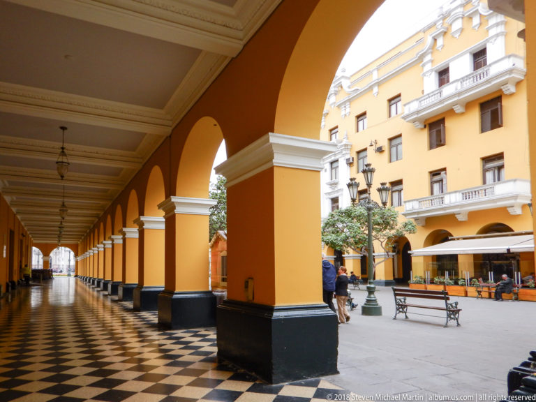 Yellow arcaded corridor with white capitals and checkerboard floor opening onto a courtyard with benches and surrounding buildings.