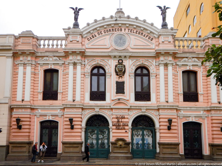 Pink-and-cream Casa de Correos y Telégrafos façade with arched entrances, pilasters, balustrade, roofline sculpture, and pedestrians.